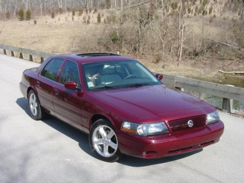 1998 Ford Crown Victoria - A maroon/dark red four-door sedan photographed from the front three-quarter angle, parked on a paved road with a guardrail and dormant wint…