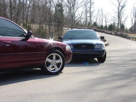1998 Ford Crown Victoria - Two Ford Crown Victoria sedans parked nose-to-nose on a paved road: a burgundy car in the foreground wearing chrome five-spoke alloy wheels…
