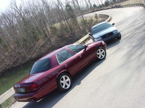 1998 Ford Crown Victoria - Elevated view of two Panther-body sedans parked nose-to-nose on a curving road beside a pond in leafless woods: a dark red Mercury sedan wi…