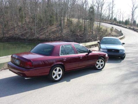 1998 Ford Crown Victoria - Two full-size sedans parked nose-to-nose on a rural road beside a pond, shot from a high angle. In front is a burgundy Mercury sedan with c…