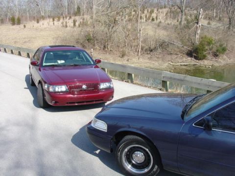 1998 Ford Crown Victoria - A dark blue/purple sedan is parked in the foreground with its front end and wheel visible, facing off with a maroon Mercury sedan parked on…