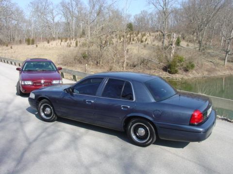 1998 Ford Crown Victoria - A dark blue/gray four-door sedan is parked on a paved road, viewed from a rear three-quarter angle, with a maroon sedan behind it and a woo…