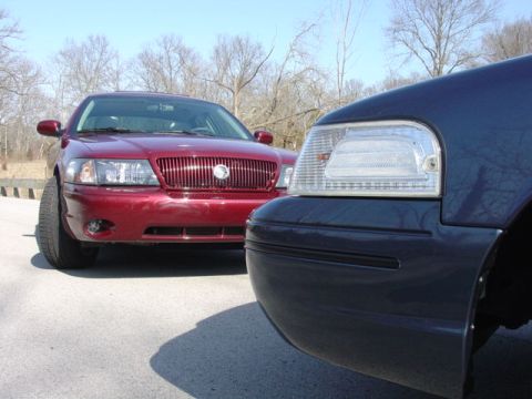 1998 Ford Crown Victoria - Two sedans nose-to-nose in a paved lot with bare trees behind; a maroon sedan with vertical chrome grille sits at left, while a dark blue s…
