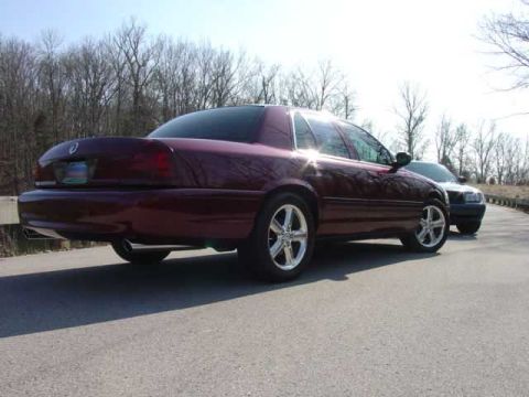 1998 Ford Crown Victoria - Rear three-quarter view of a dark burgundy Ford Crown Victoria with tinted taillights, a round emblem centered on the trunk lid, chrome fiv…