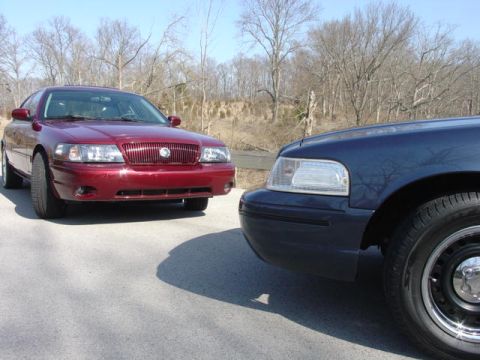 1998 Ford Crown Victoria - A burgundy Mercury full-size sedan with a body-color vertical-bar grille parked facing a dark blue Ford Crown Victoria, whose front fender…