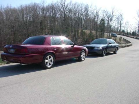 1998 Ford Crown Victoria - Two full-size sedans parked nose-to-tail on a paved curving road with bare winter trees behind; a maroon/dark-red sedan with aftermarket po…