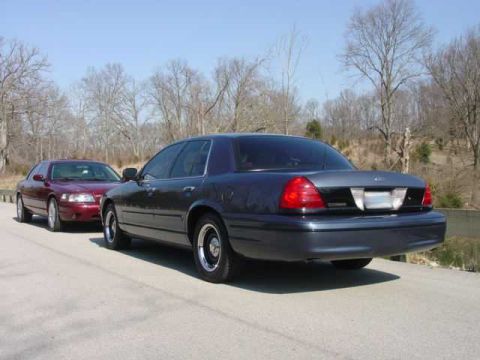 1998 Ford Crown Victoria - A dark blue/charcoal full-size Ford sedan photographed from the rear three-quarter angle on a paved road, with chrome-style wheels and visi…