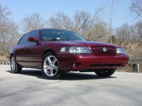 1998 Ford Crown Victoria - Dark red Mercury full-size sedan shot from the front three-quarter on a paved lot, with a chrome waterfall grille, body-colored bumpers, an…