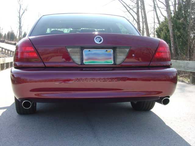 Rear of maroon Mercury sedan with dual exhaust tips, parked near wooden fence