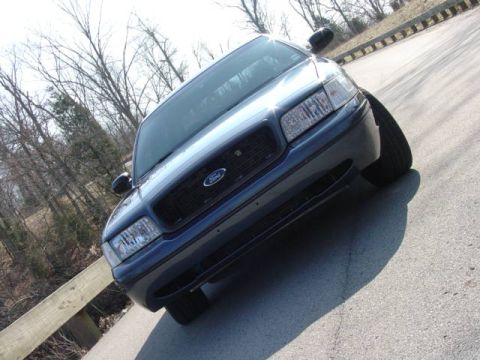 1998 Ford Crown Victoria - Dramatic low tilted angle of the front of a blue Ford Crown Victoria with black lower trim, parked on pavement beside a guardrail and bare…
