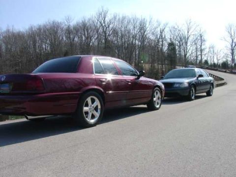 1998 Ford Crown Victoria - Maroon full-size sedan shown from the rear three-quarter with polished chrome wheels, parked on a road beside a black full-size sedan under…