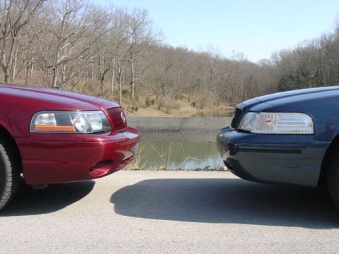 1998 Ford Crown Victoria - Two full-size sedans parked nose to nose with front bumpers nearly touching, a maroon car on the left and a dark blue car on the right, in…