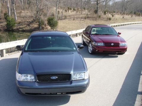 1998 Ford Crown Victoria - Elevated three-quarter front view of a dark blue-gray Ford Crown Victoria sedan with blacked-out grille, body-color bumpers and steel-wheel…