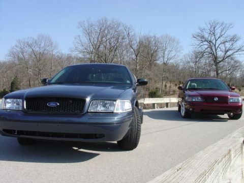 1998 Ford Crown Victoria - Low front view of a slate-gray Ford Crown Victoria with a black mesh grille parked on a bridge, with a dark red Mercury sedan parked nose-o…