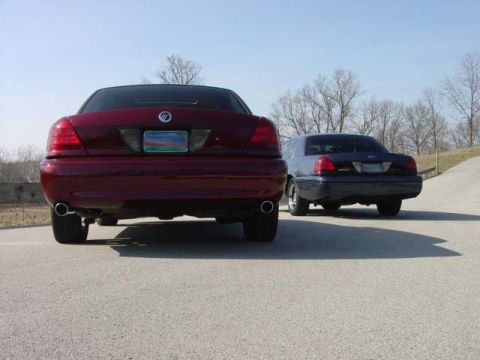 1998 Ford Crown Victoria - Head-on rear view of a dark red Mercury Grand Marquis with dual chrome exhaust tips and a rear deck spoiler, parked on pavement with a dark…