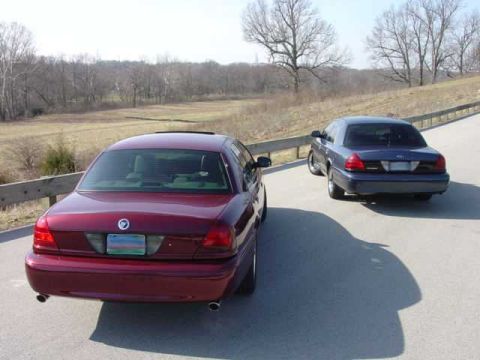 1998 Ford Crown Victoria - Elevated rear three-quarter view of a burgundy full-size sedan with a Mercury emblem and dual exhaust tips, parked on a rural road with a d…