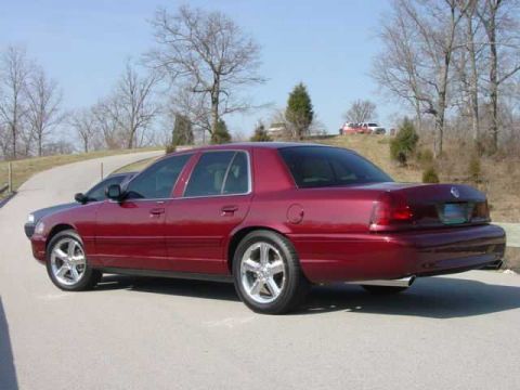 1998 Ford Crown Victoria - Rear three-quarter view of a dark red Panther-platform Mercury full-size sedan with polished five-spoke wheels and blacked-out window trim,…