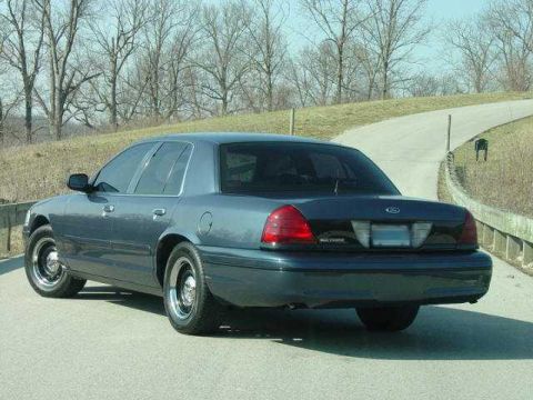 1998 Ford Crown Victoria - A dark blue/gray Ford Crown Victoria full-size sedan viewed from the rear driver's-side three-quarter angle, parked on a paved road with a…