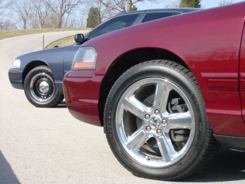 1998 Ford Crown Victoria - Low-angle close-up of the front corners of two full-size sedans lined up on pavement: a dark burgundy car in the foreground wearing polishe…
