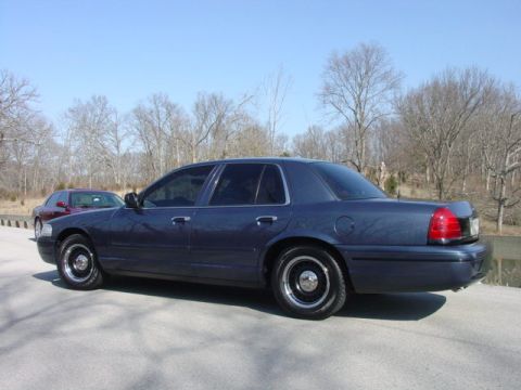 1998 Ford Crown Victoria - Dark blue Ford Crown Victoria sedan photographed from the rear three-quarter on a paved lot with bare winter trees behind, wearing black st…