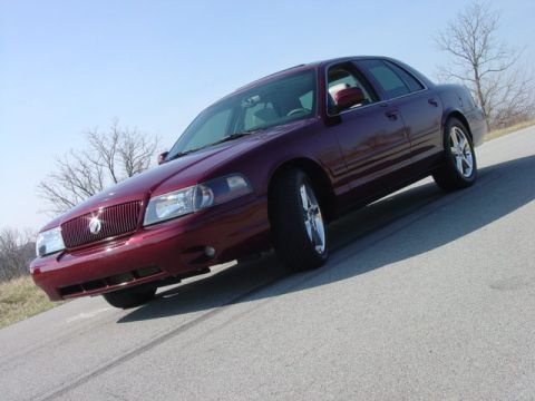 1998 Ford Crown Victoria - Low 3/4-front shot of a dark red metallic full-size sedan on a rural road, showing a body-color mesh grille with a small center emblem, cle…