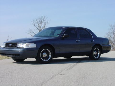 1998 Ford Crown Victoria - A dark blue Ford Crown Victoria full-size sedan shown from a 3/4 front angle, parked on an asphalt road with steel wheels and hubcaps. The…