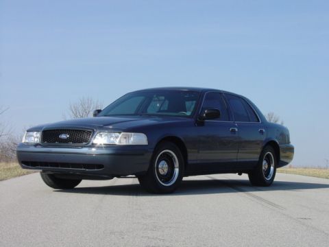 1998 Ford Crown Victoria - Low front three-quarter view of a dark slate-blue Ford Crown Victoria sedan parked on an open rural road under clear sky. The car has a bla…