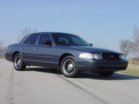 1998 Ford Crown Victoria - Three-quarter front view of a dark blue-gray Ford Crown Victoria sedan sitting on an empty two-lane road under a clear sky. It wears the bl…