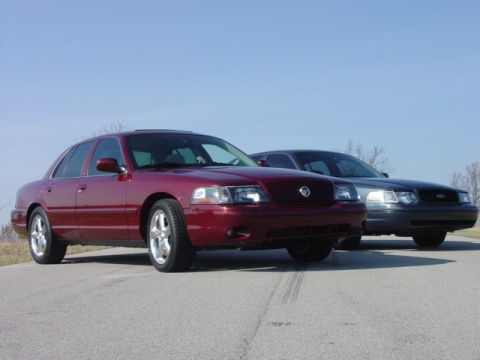 1998 Ford Crown Victoria - Two full-size four-door sedans posed nose-out on an empty asphalt road, a dark red car in front on polished multi-spoke alloy wheels with a…