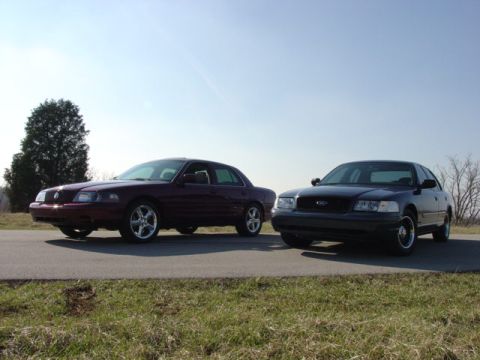 1998 Ford Crown Victoria - Two full-size sedans posed nose-out side by side on a rural blacktop under a clear sky, a dark maroon Mercury Marauder on polished five-spo…