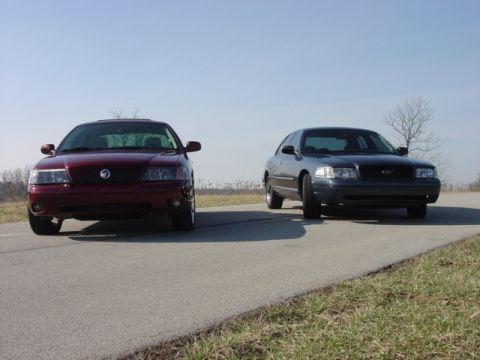 1998 Ford Crown Victoria - A dark red Mercury Marauder-style sedan with a chrome waterfall grille sits head-on beside a slate-blue Ford Crown Victoria with a dark mes…