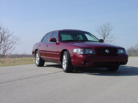 1998 Ford Crown Victoria - A deep red Mercury Marauder full-size sedan photographed from the front three-quarter on an empty rural blacktop, showing its blacked-out w…