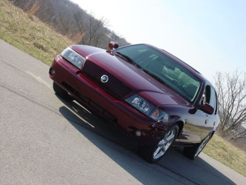1998 Ford Crown Victoria - A dark maroon/burgundy four-door sedan photographed from a low front three-quarter angle on a paved road, showing a chrome waterfall-style…