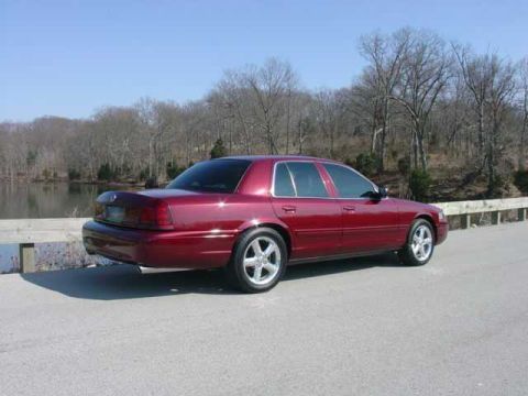 1998 Ford Crown Victoria - A dark maroon/burgundy four-door sedan photographed from the rear three-quarter angle, parked on a paved bridge or road with chrome-style m…