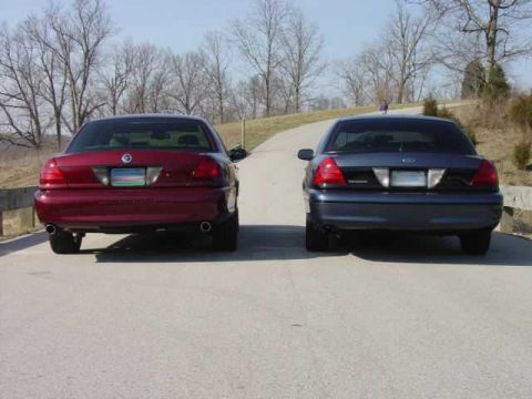 1998 Ford Crown Victoria - Two full-size sedans, a red Mercury Marauder and a dark blue Ford Crown Victoria, sit rear-to-camera side by side on a paved road.