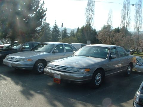1999 Ford Crown Victoria - Two Ford Crown Victorias are parked side-by-side in an outdoor lot, with trees and hills in the background.