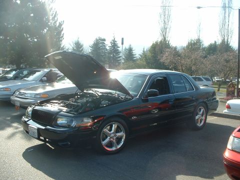 1999 Ford Crown Victoria - A dark 1999 Ford Crown Victoria is parked with its hood open, alongside other vehicles.