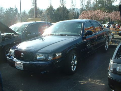 1999 Ford Crown Victoria - A dark blue 1999 Ford Crown Victoria is parked at an outdoor event, showing its front three-quarter view and chrome wheels.