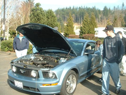 1999 Ford Crown Victoria - Light blue Ford Mustang with hood open, engine visible, with people gathered around.