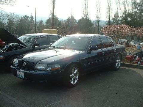 1999 Ford Crown Victoria - Two sedans parked in a lot with trees and a picnic area in the background.