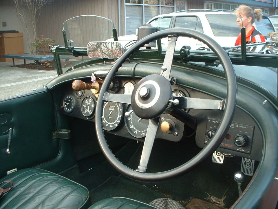 Interior of a vintage car with a large steering wheel, multiple gauges, and green leather seats. A woman is visible in the background