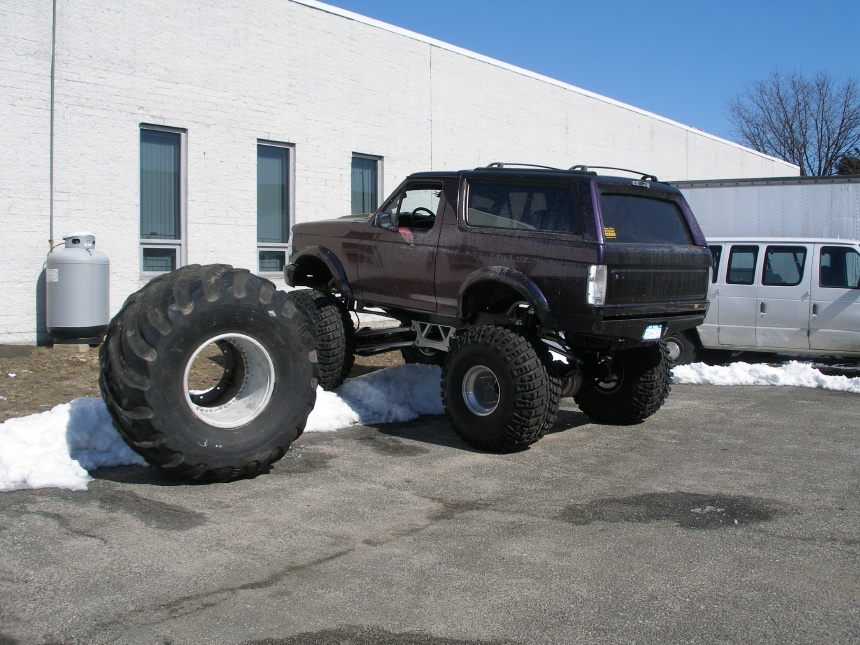 Purple lifted Ford Bronco in side view next to a huge monster truck tire in a snowy lot
