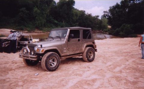 1989 Ford Bronco - A silver Jeep Wrangler sits on a sandy riverbank next to a charred vehicle.