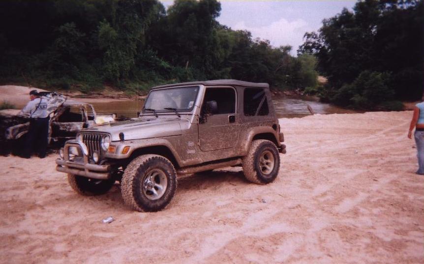 Silver Jeep Wrangler on sandy ground near a river, with a burnt vehicle in the background