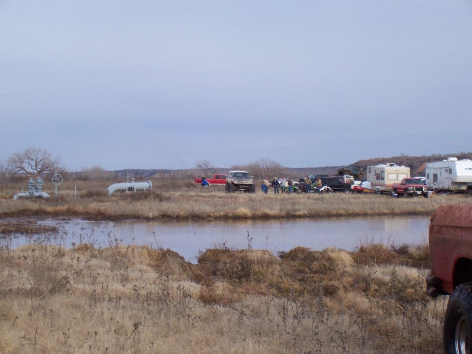 Several off-road trucks, campers, and people gathered near a muddy area with industrial pipes in the background