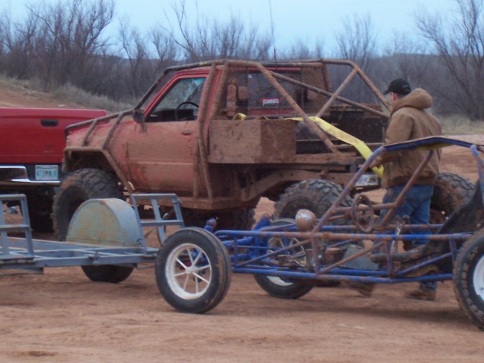 Red off-road truck covered in mud, with a custom roll cage, next to a blue buggy and a trailer