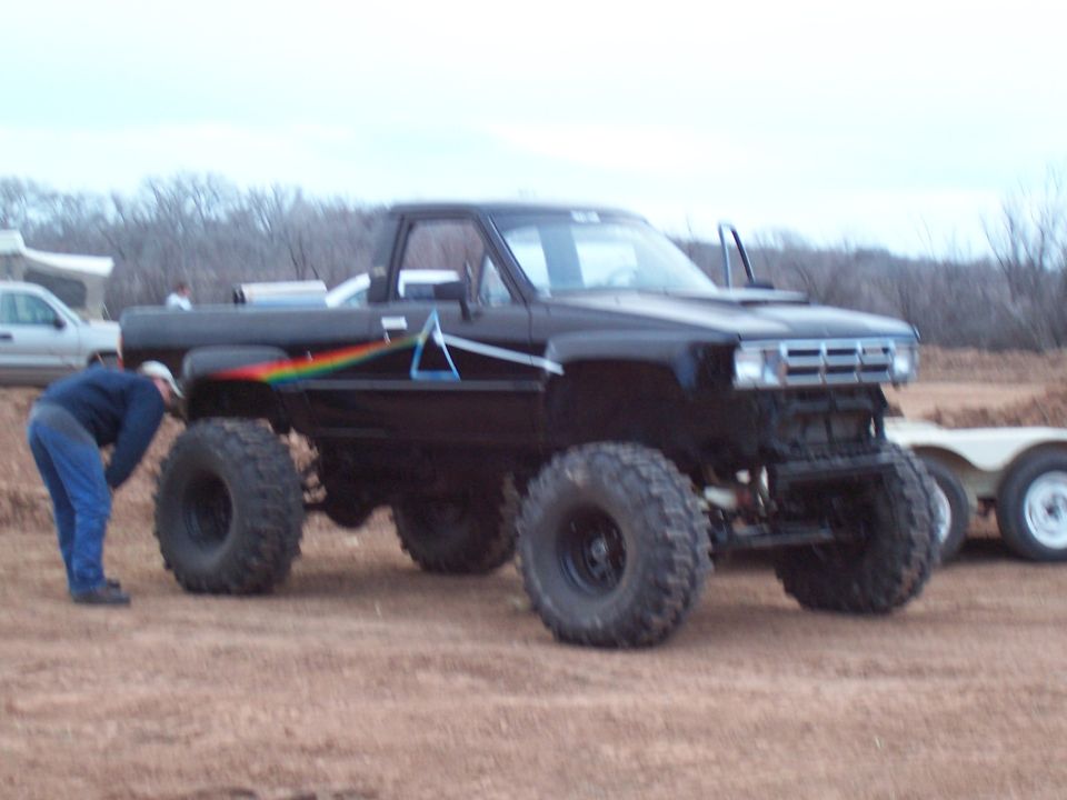 Black lifted pickup truck with Pink Floyd prism decal on the side. A person is looking at the front tire. Another vehicle and trailer are in the background
