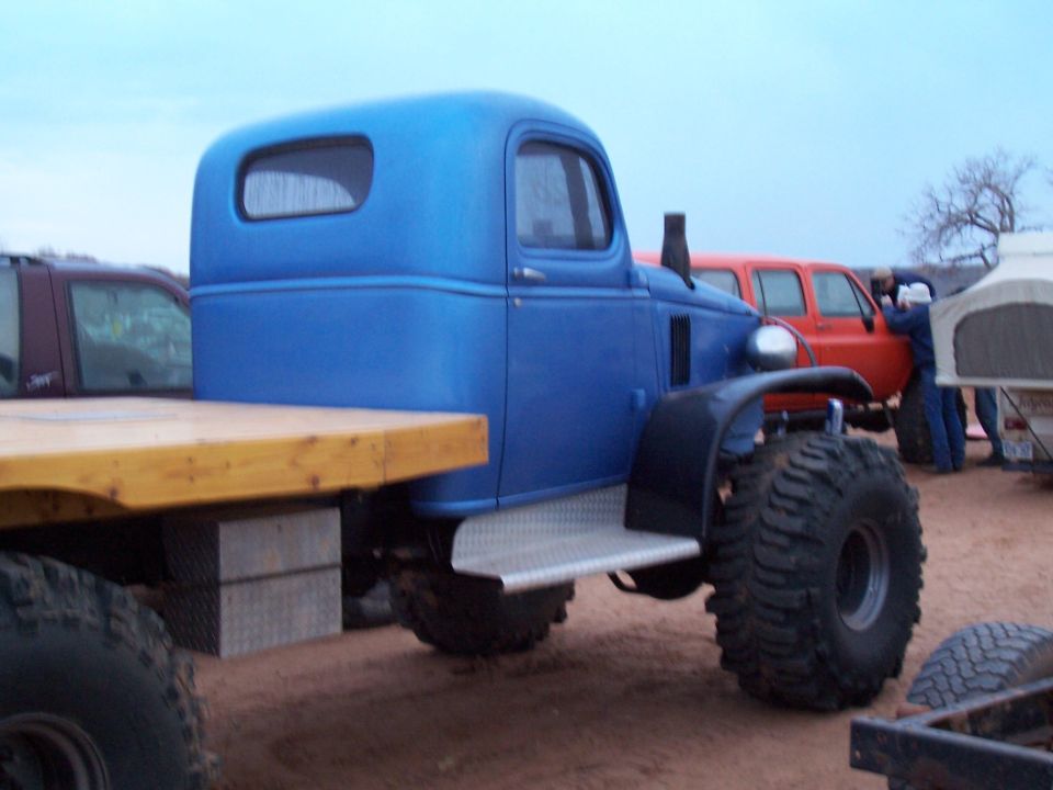 Blue flatbed truck with large off-road tires, wooden bed, and a metal step