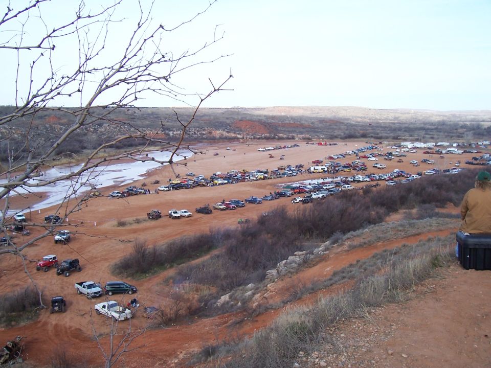 Many off-road vehicles parked in a wide, sandy area with a person sitting on a case in the foreground