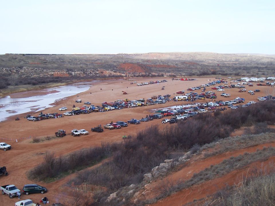 Many trucks and cars parked on a sandy riverbank with hills in the background